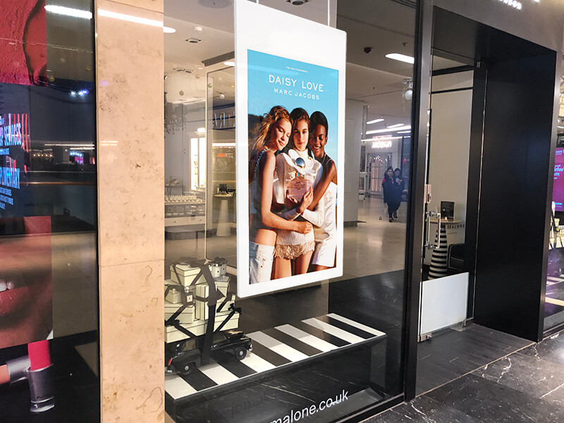 L'image représente un écran suspendu double-face d'affichage dynamique à l'intérieure d'une vitrine d'une arcade commerciale. L'écran affiche une publicité pour un article du magasin.