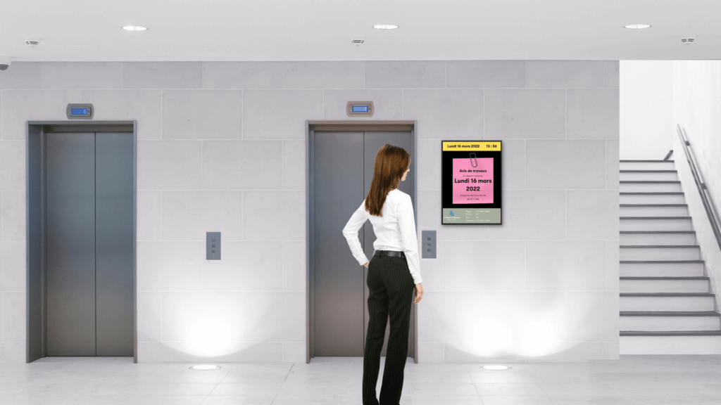 L'image représente une allée d'immeuble équipée d'un écran d'information digital placé à coté de l'ascenseur. L'écran d'accueil digital affiche les informations importantes diffusées par la régie immobilière de l'immeuble.